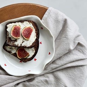 May include: Two slices of dark bread topped with cream cheese and fig slices on a white plate with red heart accents. The plate sits on a wooden surface with a gray linen cloth.