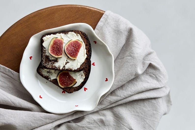 May include: Two slices of dark bread topped with cream cheese and fig slices on a white plate with red heart accents. The plate sits on a wooden surface with a gray linen cloth.