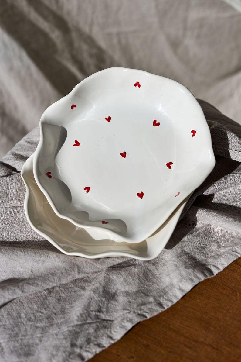 May include: Two white ceramic plates with wavy edges, each adorned with small red heart designs. The plates are stacked on top of each other, creating a layered effect. The plates are on a gray linen cloth.