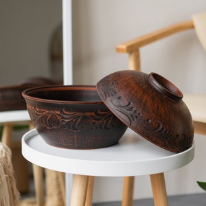 May include: A set of two brown ceramic bowls with intricate carved designs. The bowls are sitting on a white table with wooden legs.