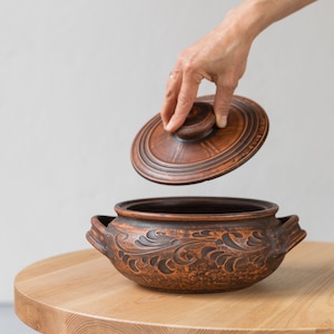 May include: A brown ceramic pot with a lid and intricate floral designs. The pot is sitting on a wooden table.