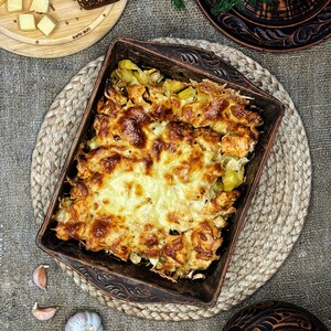 May include: A rectangular brown ceramic baking dish filled with a baked casserole of golden-brown melted cheese, chicken, and potatoes, featuring decorative embossed patterns on the rim and handles, presented in a rustic kitchen setting.