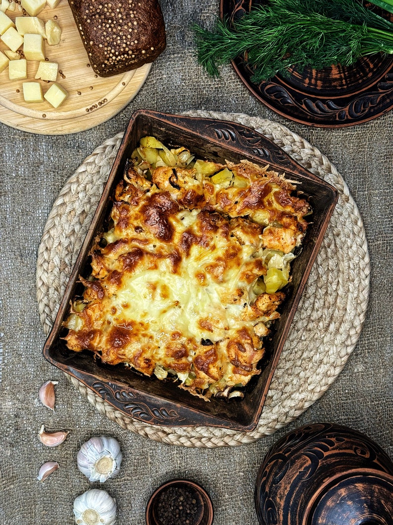 May include: A rectangular brown ceramic baking dish filled with a baked casserole of golden-brown melted cheese, chicken, and potatoes, featuring decorative embossed patterns on the rim and handles, presented in a rustic kitchen setting.