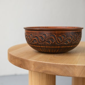 May include: A brown ceramic bowl with a decorative black floral design sits on a light wood table with rounded legs.