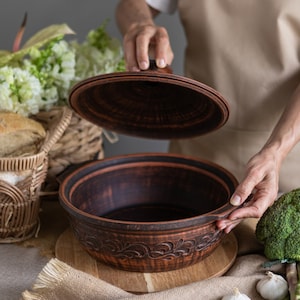 May include: A brown ceramic pot with a lid, decorated with a floral design. The pot is sitting on a wooden cutting board, surrounded by garlic cloves and asparagus spears.