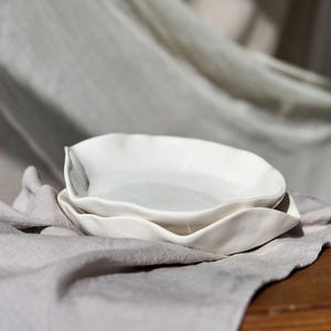 May include: Two white ceramic dishes with a wavy, organic shape are stacked on a gray linen cloth. The dishes have a smooth, glossy finish and are set against a neutral background, suggesting a minimalist aesthetic.