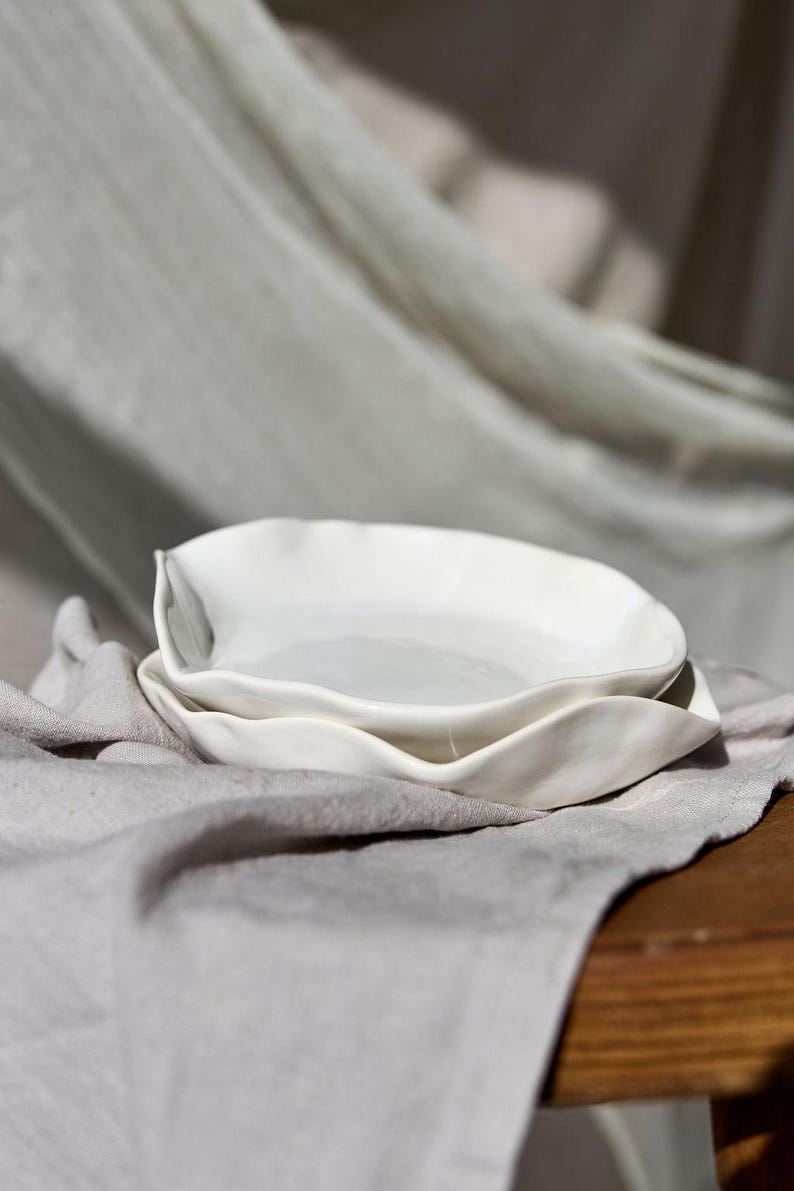 May include: Two white ceramic dishes with a wavy, organic shape are stacked on a gray linen cloth. The dishes have a smooth, glossy finish and are set against a neutral background, suggesting a minimalist aesthetic.