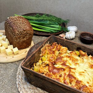May include: A rectangular brown ceramic baking dish filled with a golden-brown baked potato and cheese casserole, accompanied by a loaf of dark rye bread topped with coriander seeds, cubed cheese, fresh green onions, dill, and garlic bulbs.