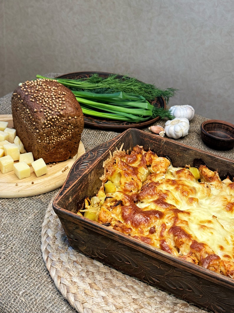 May include: A rectangular brown ceramic baking dish filled with a golden-brown baked potato and cheese casserole, accompanied by a loaf of dark rye bread topped with coriander seeds, cubed cheese, fresh green onions, dill, and garlic bulbs.