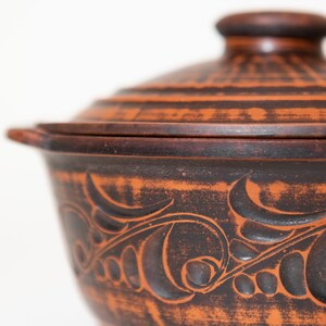 Może przedstawiać: Rustykalne, terakotowe ceramiczne naczynie do zapiekania z pokrywką. Naczynie posiada ciemnobrązowy, bogaty kwiatowy wz&oacute;r wyrzeźbiony na powierzchni. Pokrywka ma mały, zaokrąglony uchwyt. Naczynie przeznaczone jest do gotowania i serwowania.