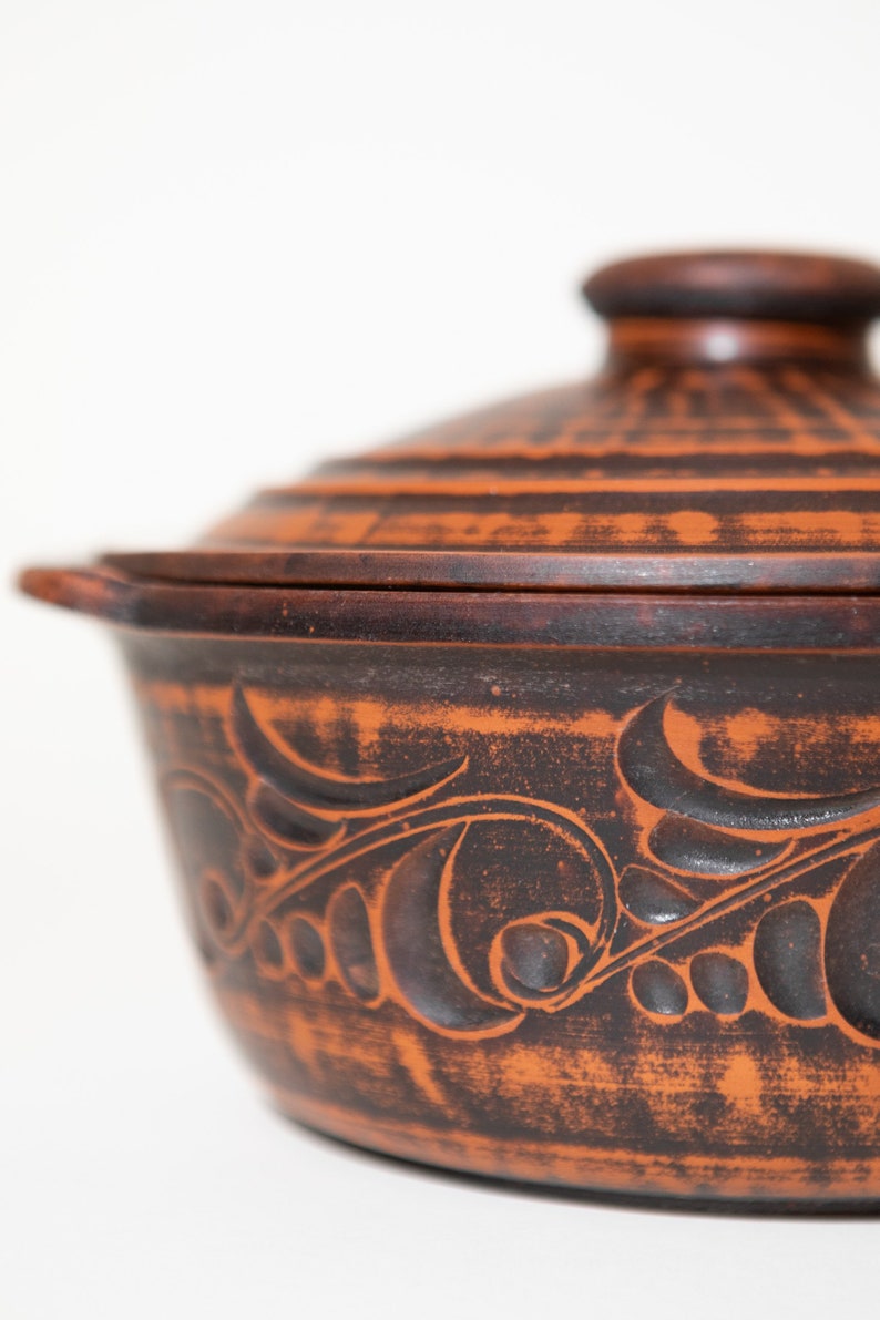 Może przedstawiać: Rustykalne, terakotowe ceramiczne naczynie do zapiekania z pokrywką. Naczynie posiada ciemnobrązowy, bogaty kwiatowy wz&oacute;r wyrzeźbiony na powierzchni. Pokrywka ma mały, zaokrąglony uchwyt. Naczynie przeznaczone jest do gotowania i serwowania.