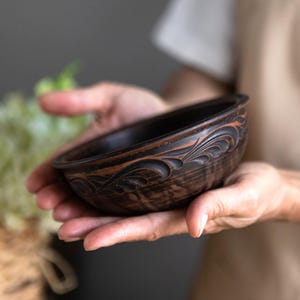 Puede incluir: Un cuenco de cerámica marrón oscuro con intrincados diseños tallados. El cuenco se sostiene en las manos de una persona.