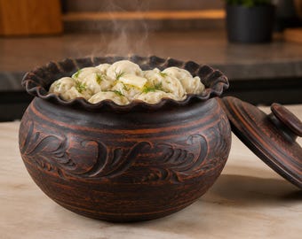 May include: A dark brown clay pot with a ruffled rim and embossed swirling patterns, filled with steaming dumplings topped with a pat of butter and fresh green dill. A matching clay lid with a small handle rests beside the pot.