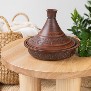 Puede incluir: Una tagine de cer&aacute;mica marr&oacute;n con intrincados dise&ntilde;os florales se encuentra sobre una mesa de madera clara con tres patas. La tagine es una olla de cocci&oacute;n tradicional marroqu&iacute; con una tapa c&oacute;nica.