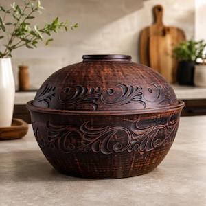May include: A dark brown clay cooking pot with a matching lid, featuring a rounded shape and intricate carved swirling patterns along the exterior surface. The vessel has a wide base and a smooth, textured finish consistent with natural earthenware.