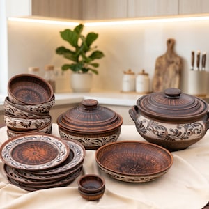 May include: A collection of brown clay dinnerware featuring dark brown and cream-coloured floral patterns. The set includes a large lidded pot with handles, a lidded casserole dish, several stacked bowls, flat plates, and two small individual dipping bowls.