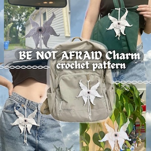 Op de afbeelding: Witte gehaakte hanger in de vorm van een vlinder met een zwart oog in het midden. De hanger hangt aan een rugzak, een autoruitspiegel en een plant. De tekst "BE NOT AFRAID Charm crochet pattern" is te zien op de afbeelding.