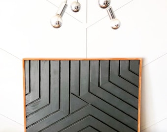 May include: A framed geometric wall art piece with a black wood design and a natural wood frame. The art piece is hanging on a white wall with a modern light fixture above it.
