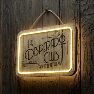 May include: A rectangular wooden sign hanging by a rope, featuring a glowing yellow LED border. The sign displays the text 'THE DESPERADO CLUB' and 'SO FUN IT HURTS' in black lettering, accompanied by a graphic illustration of a vertical sword.