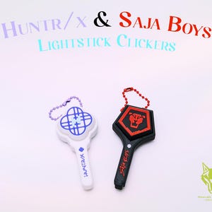 May include: Two lightstick clickers, one white and one black, with keychains. The white clicker has a blue design and the text "HUNTR/X". The black clicker has a red and black design and the text "Saja Boys". The text "HUNTR/X & Saja Boys Lightstick Clickers" is above the clickers.