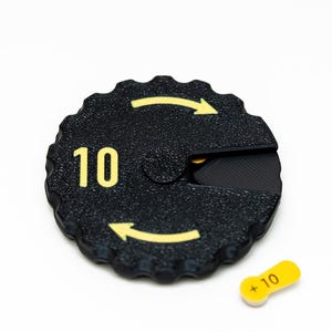 May include: Black plastic game piece with a yellow number "10" and arrows. A separate yellow plastic piece with the text " +10" is also shown.