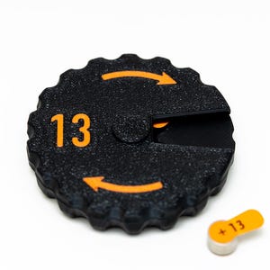 May include: A black plastic watch battery with an orange label that says "13". The battery is on a white surface.