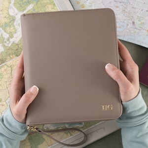 May include: A beige leather travel wallet with a zip closure and a gold handle. The wallet has the initials "H.G." embossed in gold on the front.