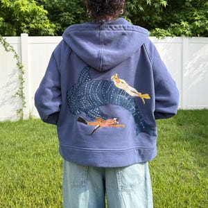 May include: A blue hooded sweatshirt with a large whale shark and two divers embroidered on the back. The sweatshirt is a solid blue color and has a drawstring hood. The divers are in yellow and orange swimsuits.