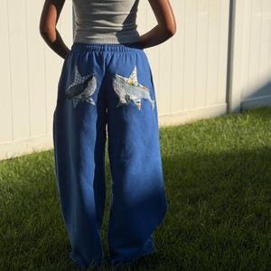 May include: Blue sweatpants featuring two large star-shaped patches on the back. Each patch displays a whale graphic layered over a patterned fabric background. The sweatpants have an elastic waistband and a loose, relaxed fit throughout the legs.