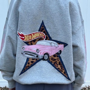 May include: Gray sweatshirt featuring a pink classic car graphic with the Hot Wheels logo. The car is set against a leopard print star. The sleeves have pink stripes.