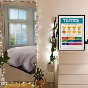 K&ouml;nnte beinhalten: Ein Ganzk&ouml;rperspiegel mit Lichterketten steht neben einem Kaktus in einem wei&szlig;en Topf. Ein gerahmtes "T&auml;gliches Emotionales Check-in-Diagramm" befindet sich auf einer wei&szlig;en Kommode. Das Diagramm hat einen blauen Hintergrund und listet Gef&uuml;hle und Bed&uuml;rfnisse auf.