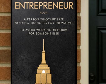May include: Black rectangular sign with gold text that reads "ENTREPRENEUR" and a definition. Below is a gold illustration of a building. The sign is mounted on a brick wall next to a black door.