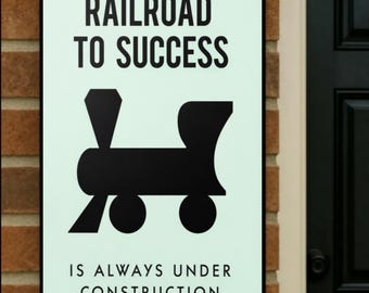 May include: A framed sign with the text "RAILROAD TO SUCCESS" above a black train graphic. Below the graphic, the text reads "IS ALWAYS UNDER CONSTRUCTION". The sign is mounted on a brick wall.