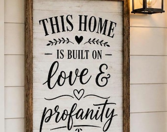 May include: A framed wooden sign with the text "THIS HOME IS BUILT ON love & profanity" in black script. The sign is mounted on a white wooden wall, with a black lantern and greenery visible.