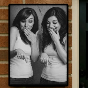 Op de afbeelding: Zwart-wit ingelijste foto van twee vrouwen. Ze dragen tanktops en jeans. Ze bedekken hun mond met hun handen en wijzen naar beneden met hun andere handen. De vrouwen lachen.