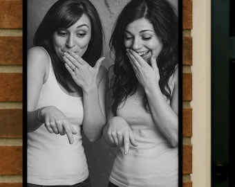 May include: Black and white framed photograph of two women. Both women are wearing tank tops and jeans. They are covering their mouths with their hands and pointing down with their other hands. The women are smiling.