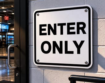 May include: A white rectangular sign with black text that reads "ENTER ONLY" is mounted on a gray brick wall. The sign has a black border and is secured with four silver screws. A glass door and the word "EXIT" are visible in the background.