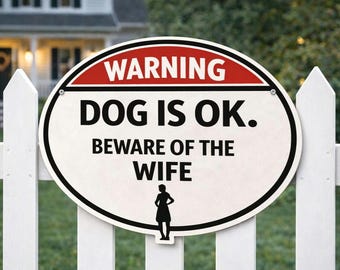 Könnte beinhalten: Ovales Schild mit rotem und weißem Rand, montiert an einem weißen Zaun. Auf dem Schild steht "WARNING DOG IS OK. BEWARE OF THE WIFE" in fetten schwarzen Buchstaben. Am unteren Rand des Schildes befindet sich eine Frauensilhouette.