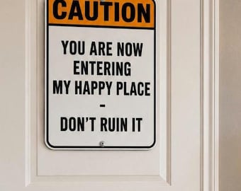 May include: A rectangular sign with the word "CAUTION" in orange letters at the top. Below, the sign reads "YOU ARE NOW ENTERING MY HAPPY PLACE - DON'T RUIN IT" in black text. The sign is mounted on a white door.