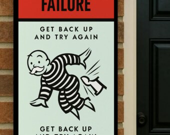 May include: A rectangular sign with a red top section that reads "FAILURE." Below, the sign features a Monopoly-inspired illustration of a figure in a striped prison uniform, with the text "GET BACK UP AND TRY AGAIN" above and below the image.