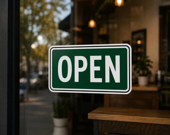 May include: A rectangular green and white "OPEN" sign is displayed on a glass door. The sign has a white border and bold white text. The background shows a blurred interior with plants and wooden surfaces.