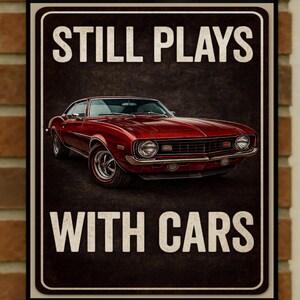 May include: A vintage-style metal sign with a red classic car illustration. The sign reads "STILL PLAYS WITH CARS" in white capital letters against a dark brown background. The car is a detailed rendering of a classic muscle car.