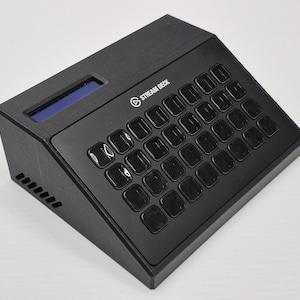 May include: Black Stream Deck with multiple square buttons and a small blue screen. The device has the Stream Deck logo on the top right. The buttons are arranged in a grid pattern.