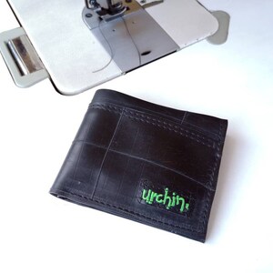 Puede incluir: Una cartera de cuero negro hecha de c&aacute;maras de bicicleta recicladas. La cartera tiene un logotipo bordado en verde que dice "urchin."