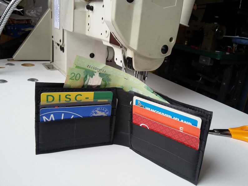 Puede incluir: Una cartera negra hecha de c&aacute;maras de aire de neum&aacute;ticos recicladas, abierta para revelar varias tarjetas y billetes canadienses. La cartera tiene una superficie texturizada y est&aacute; sobre una superficie blanca.