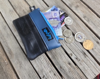 May include: A black and blue coin purse with a zip, featuring the word "urchini". The purse is open, displaying banknotes and coins. The purse is made of a textured material and rests on a wooden surface.