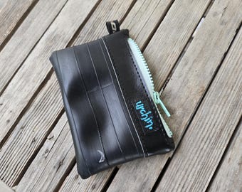 May include: A black rectangular coin purse constructed from recycled rubber inner tube material. It features a light mint green zip closure and a small black fabric tag with the word urchin embroidered in light blue thread on the front.