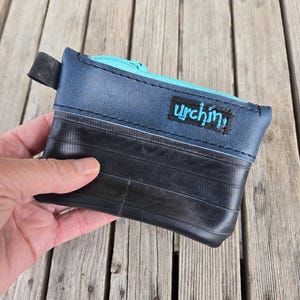 May include: A small, rectangular pouch made from recycled materials. The top section is navy blue with a teal zip, and the bottom is black. The word "urchin" is embroidered in teal on the navy blue section. A black loop is attached to the side.