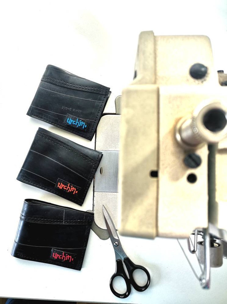 Puede incluir: Tres carteras de cuero negro con la palabra "urchin" impresa en ellas en diferentes colores. Las carteras est&aacute;n sobre una superficie blanca junto a un par de tijeras plateadas y una m&aacute;quina de coser.