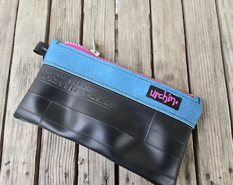 May include: A rectangular zippered pouch constructed from black rubber with embossed tyre markings. The top section features a bright blue fabric band with a pink zip and a black rectangular patch displaying the word urchin in pink text.